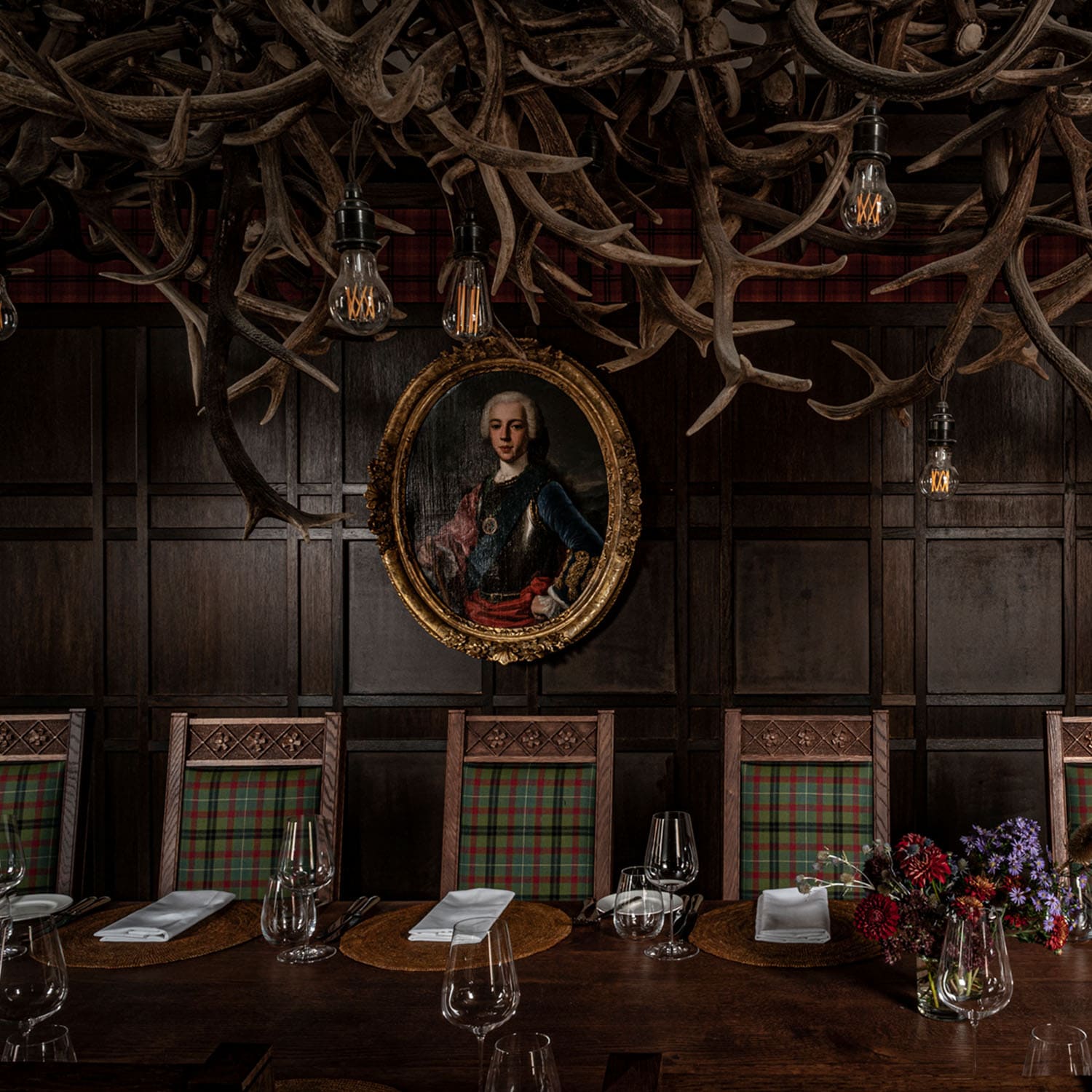 A section of the hand-carved table in The Scottish Room with oak chairs upholstered in green and red tartan, with artwork in the background of: “Prince Charles Edward Stewart (‘The Young Pretender’ (1720-1788)”,  1740 by (Giorgio) Domenico Dupra (1689–1770)